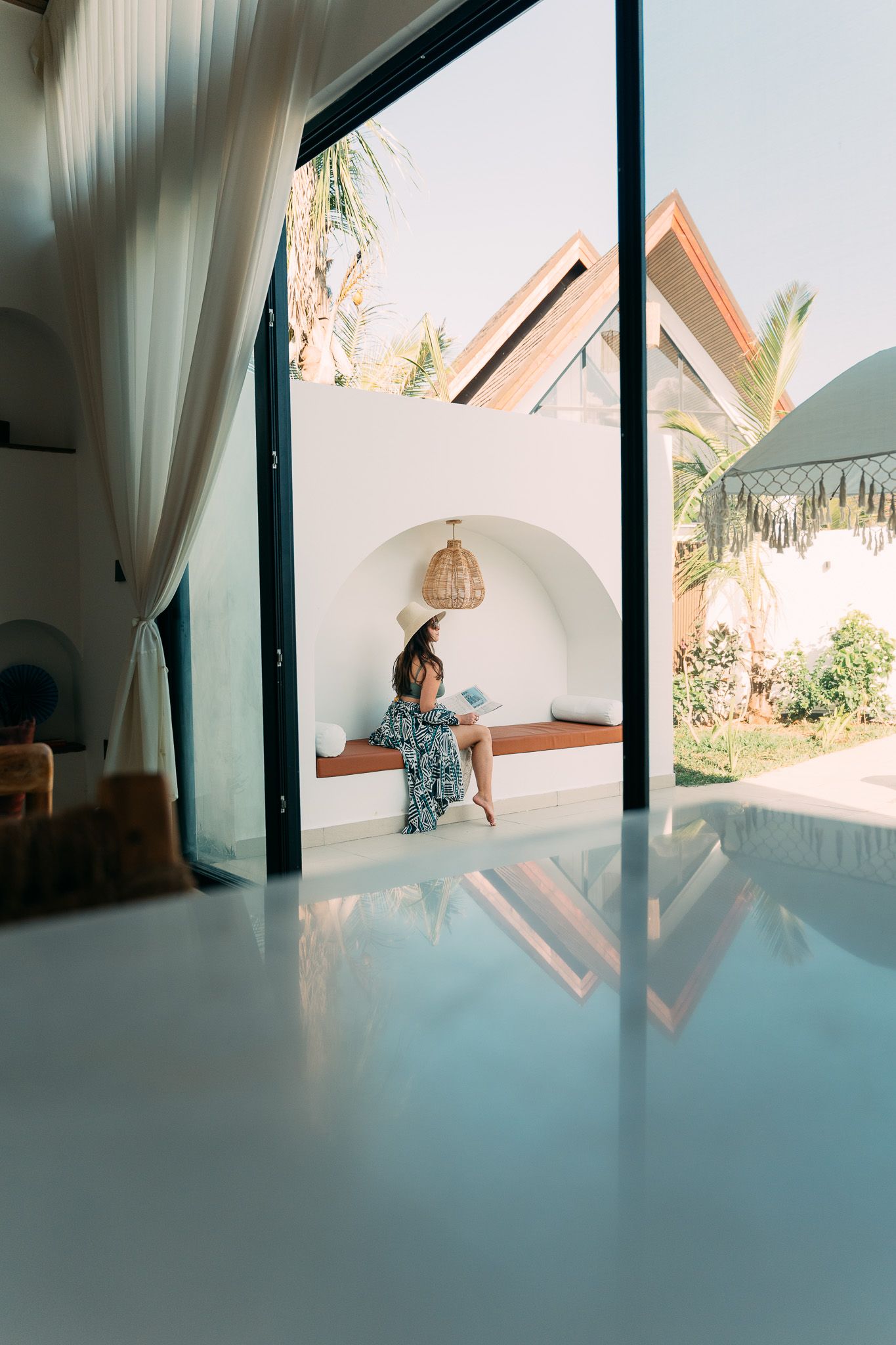 Lifestyle shot — woman reading on built-in bench with white pillows, pool and tropical garden visible — Airbnb Zanzibar photography