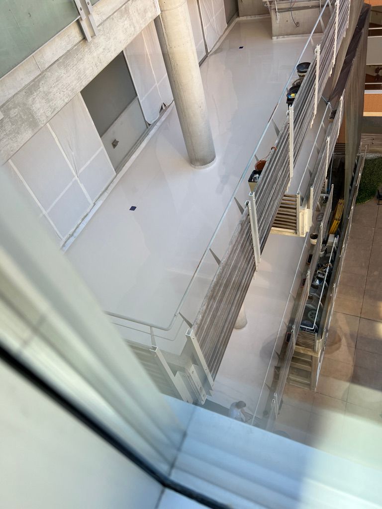 Multi-level healthcare facility interior with white seamless floors, large cylindrical column, metal railings and potted plants along walkway