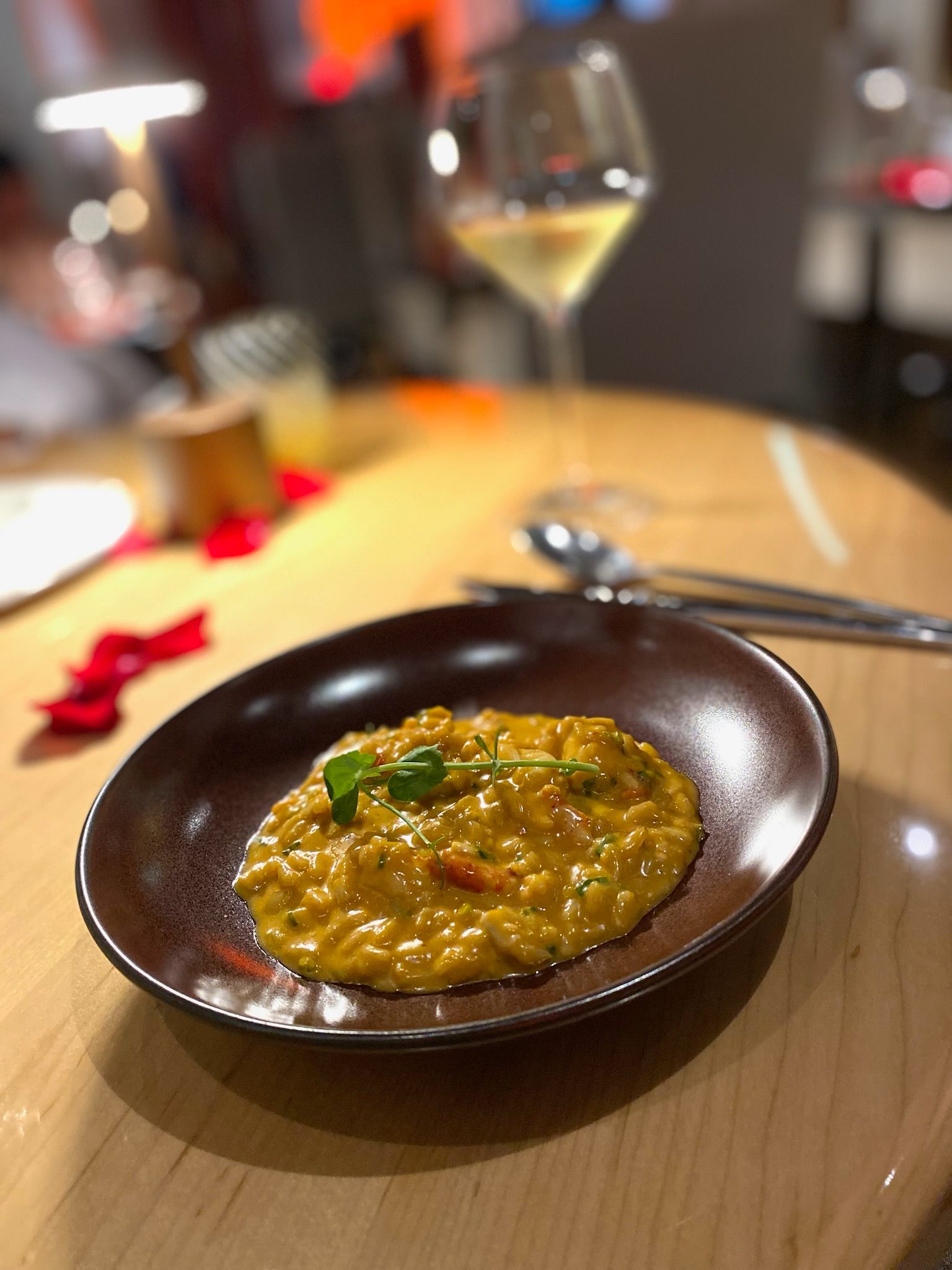 Marmalade tasting menu — golden-orange creamy dish in a shallow dark bowl with fresh herbs and wine glass, wooden table with rose petals
