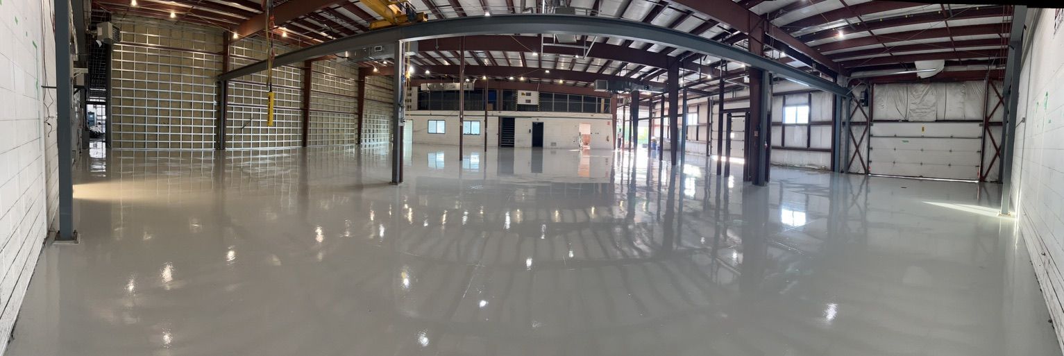 Large empty industrial warehouse with polished concrete floor, metal beams and columns, multiple overhead doors and enclosed office at far end