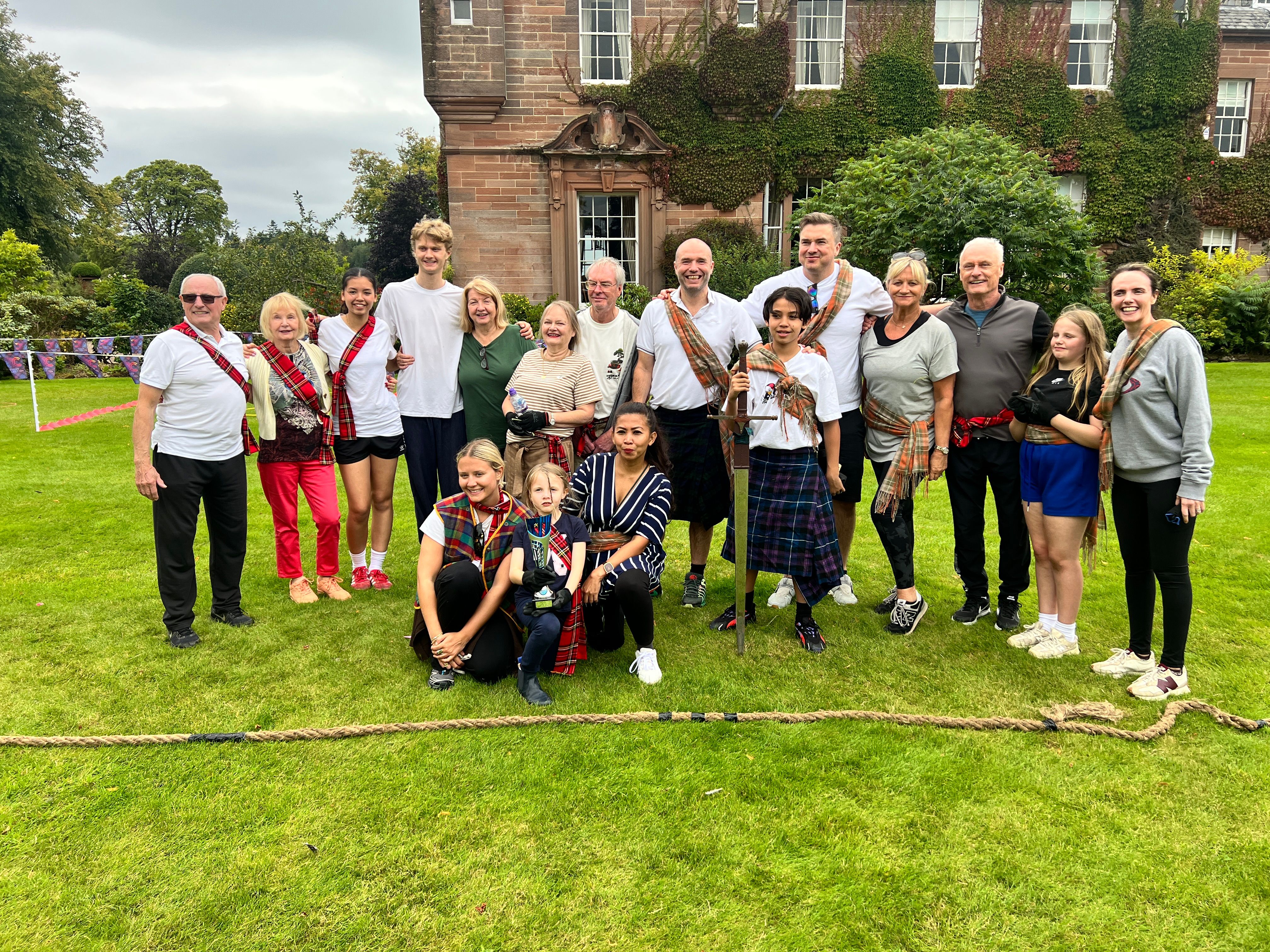 Highland Games team building — group of 17 in tartans celebrating with trophy and tug of war rope at a grand estate