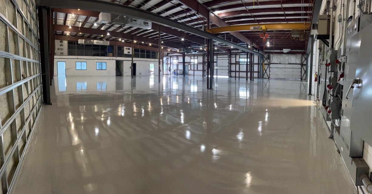 Large empty industrial warehouse with polished concrete floor reflecting ceiling lights, exposed metal beams and staircase to upper level