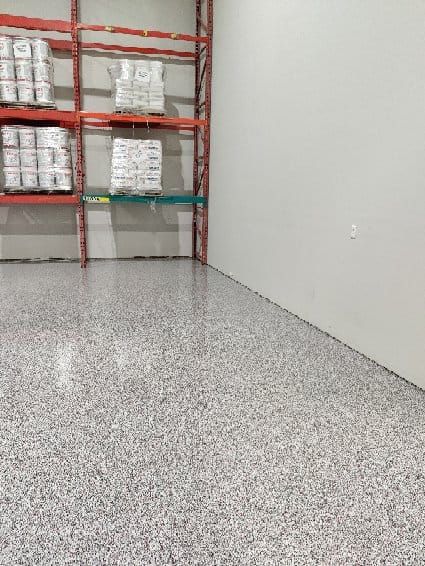 Corner of a warehouse with speckled grey epoxy floor and red metal shelving stocked with bags and containers
