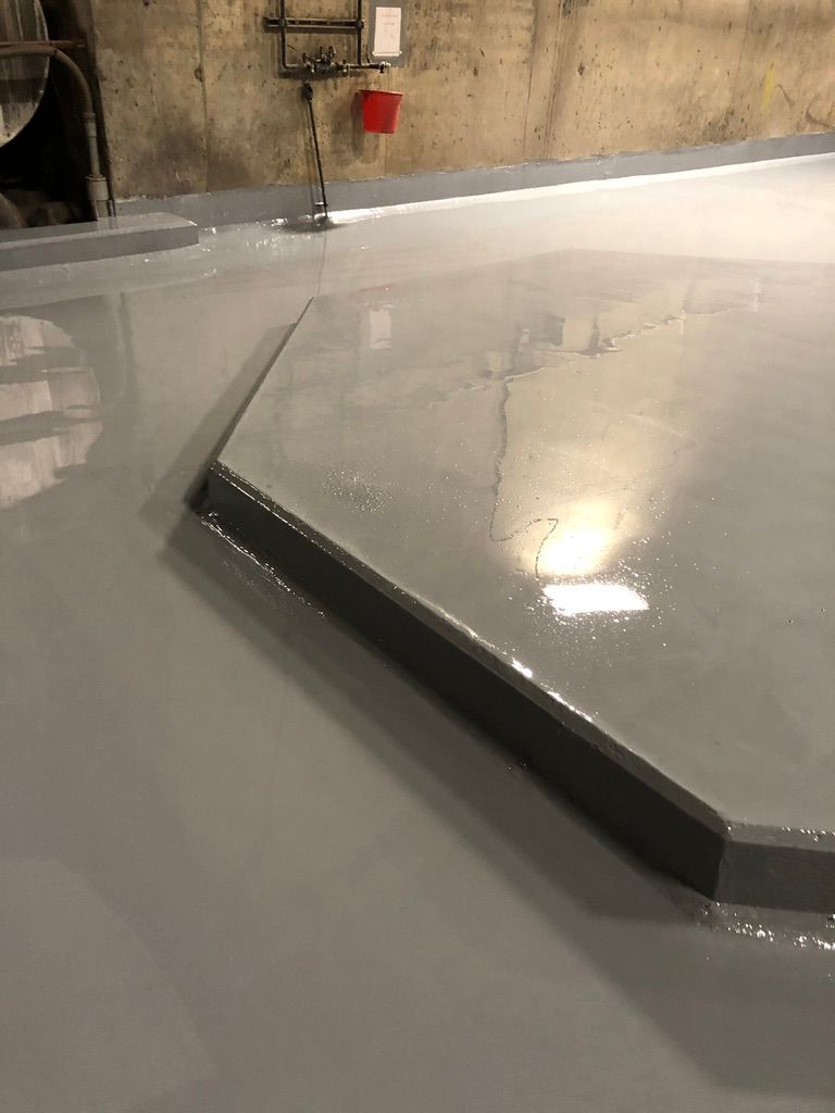 Close-up of a freshly coated smooth grey seamless floor in a wastewater facility with a raised rectangular containment platform and pipes along the wall