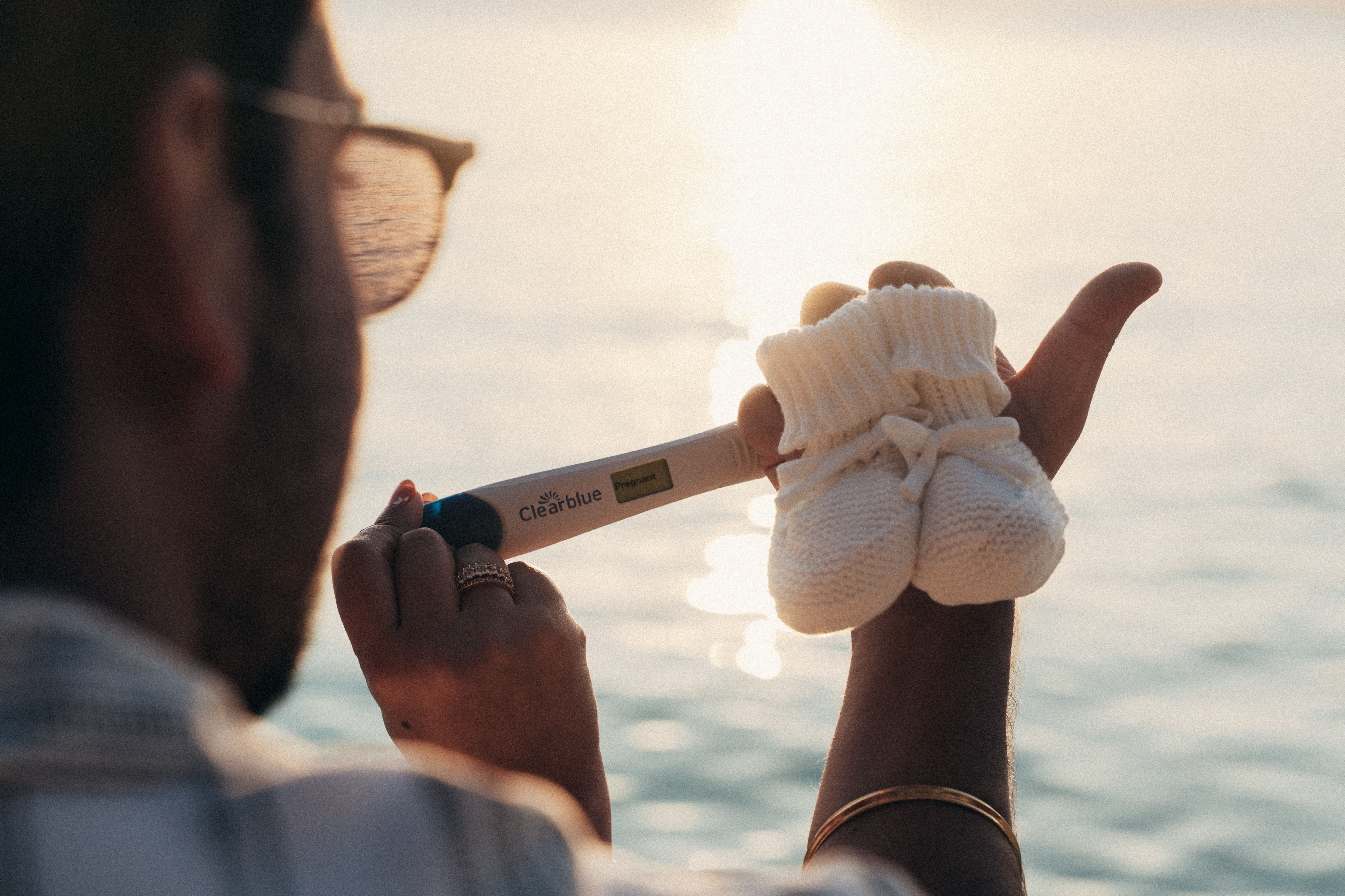 Pregnancy test and baby booties held against sunset over ocean - pregnancy announcement