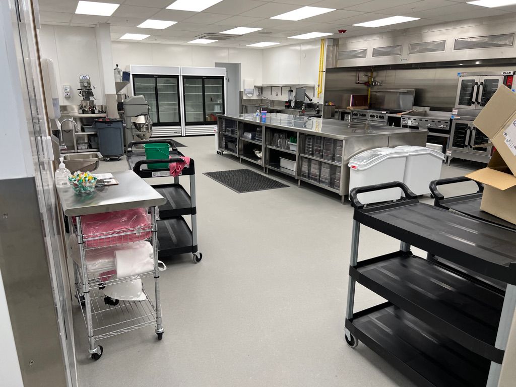 Large healthcare commercial kitchen with stainless steel counters, rolling carts, glass-door refrigerators and ovens — seamless light-colored floor with fluorescent lighting