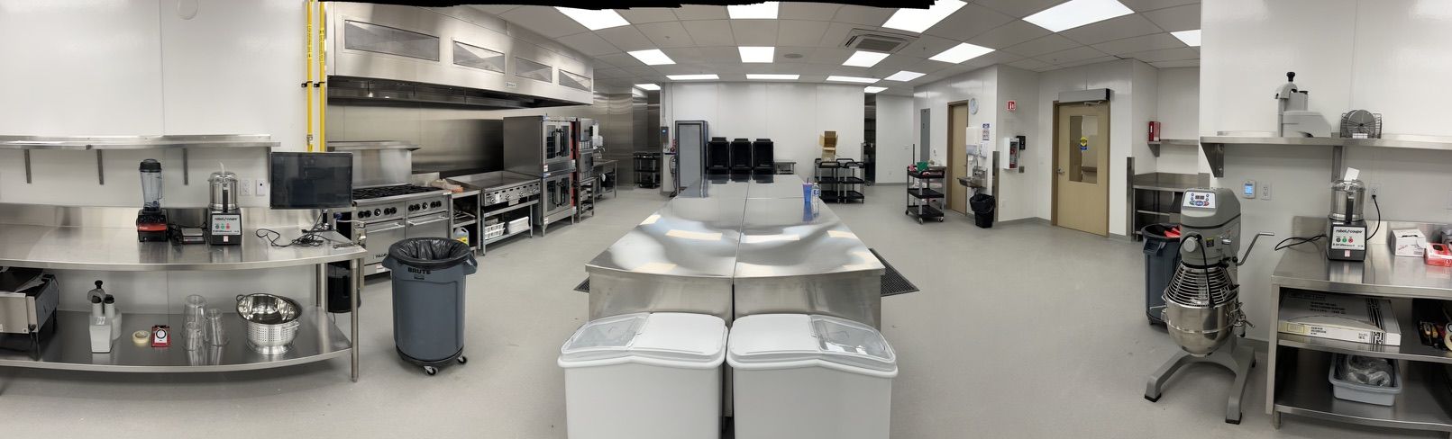 Large commercial healthcare kitchen with stainless steel counters, ovens, stoves, ventilation hoods and blenders — seamless floor and bright ceiling lighting