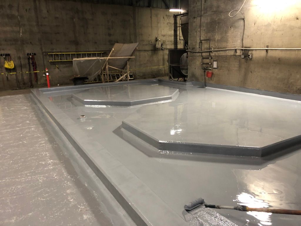 Freshly coated shiny grey seamless floor in a water treatment facility with raised platform sections, paint roller on the surface and yellow ladder against the wall