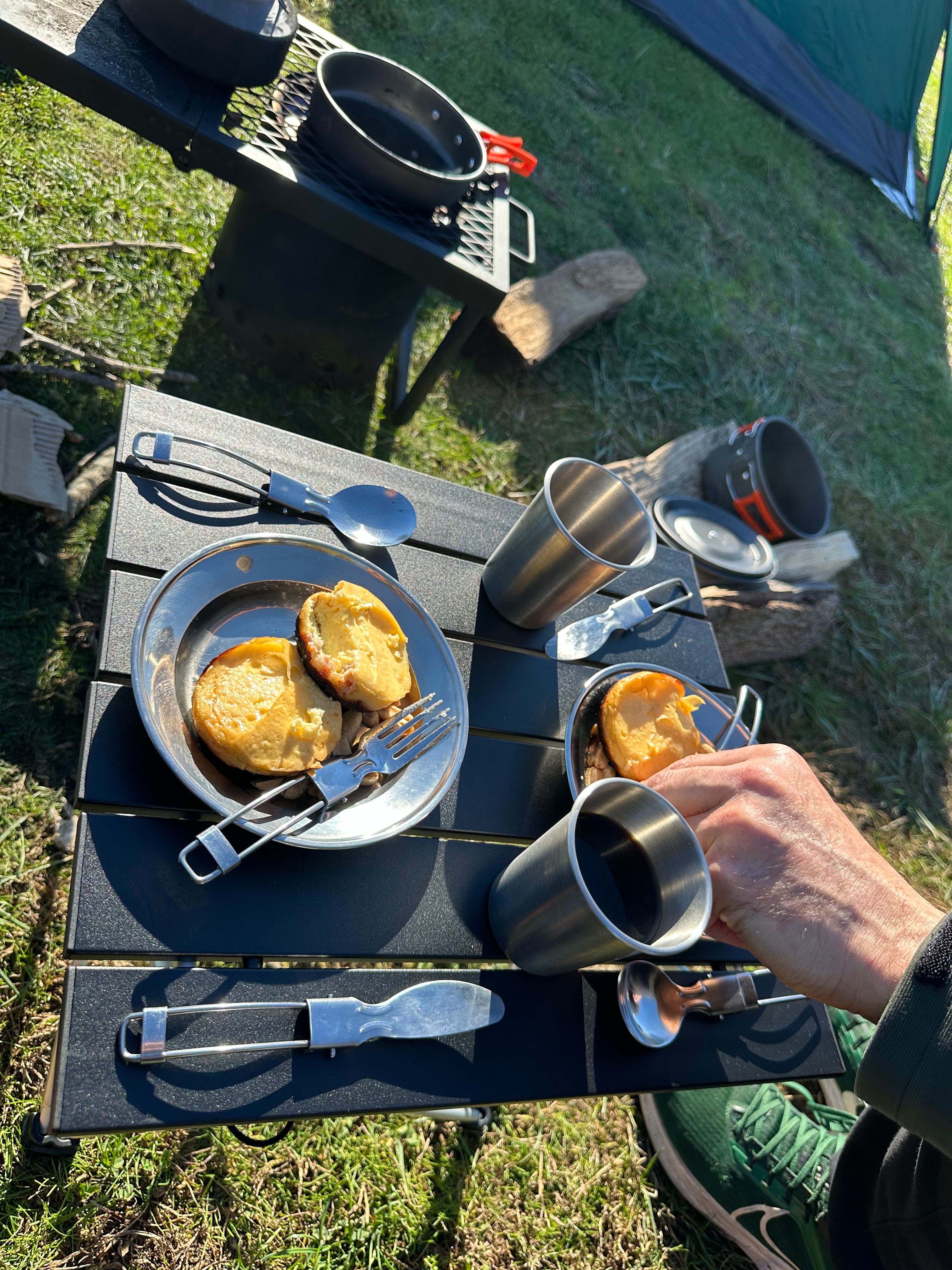 Camping breakfast setup — small black table with metal plates of food, metal cups, person in green jacket, grill and tent in background