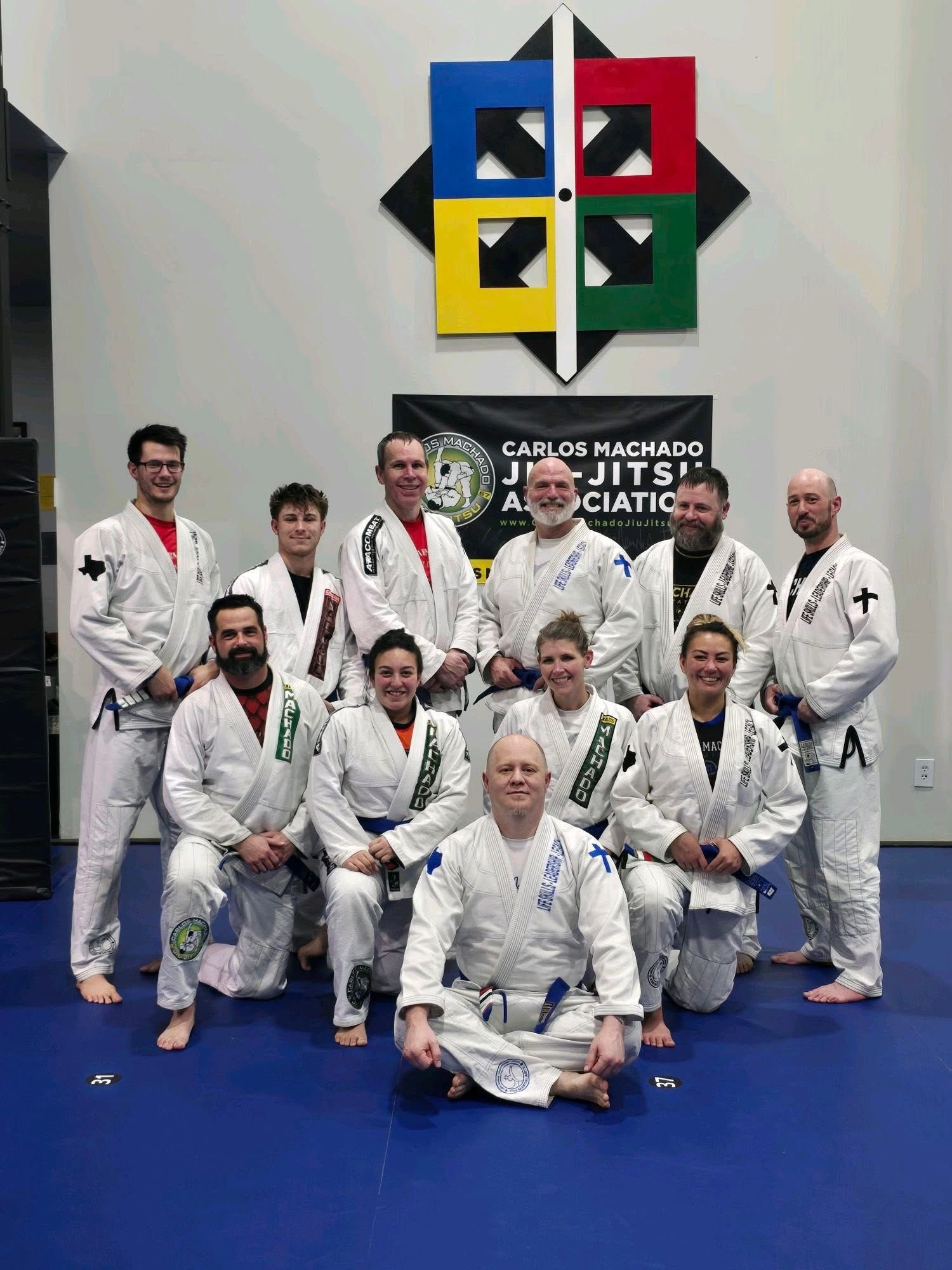 Adult Jiu-Jitsu students in gis with blue belts in front of Machado banner