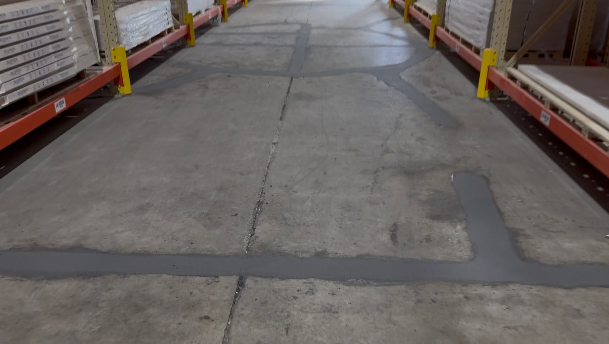 Warehouse aisle before-shot showing patched concrete floor with shelves loaded with plastic-wrapped product