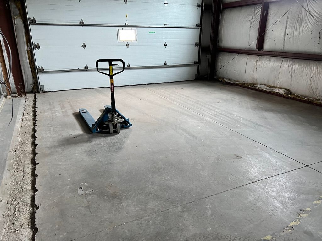 Empty indoor industrial space with concrete floor and white garage door, blue pallet jack in center and metal walls with insulation