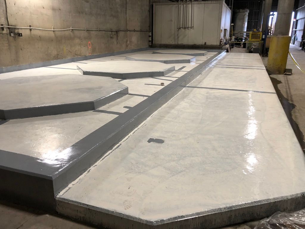 Smooth grey and white seamless floor with multiple raised hexagonal containment pads in a water treatment facility, yellow safety column and caution tape in foreground
