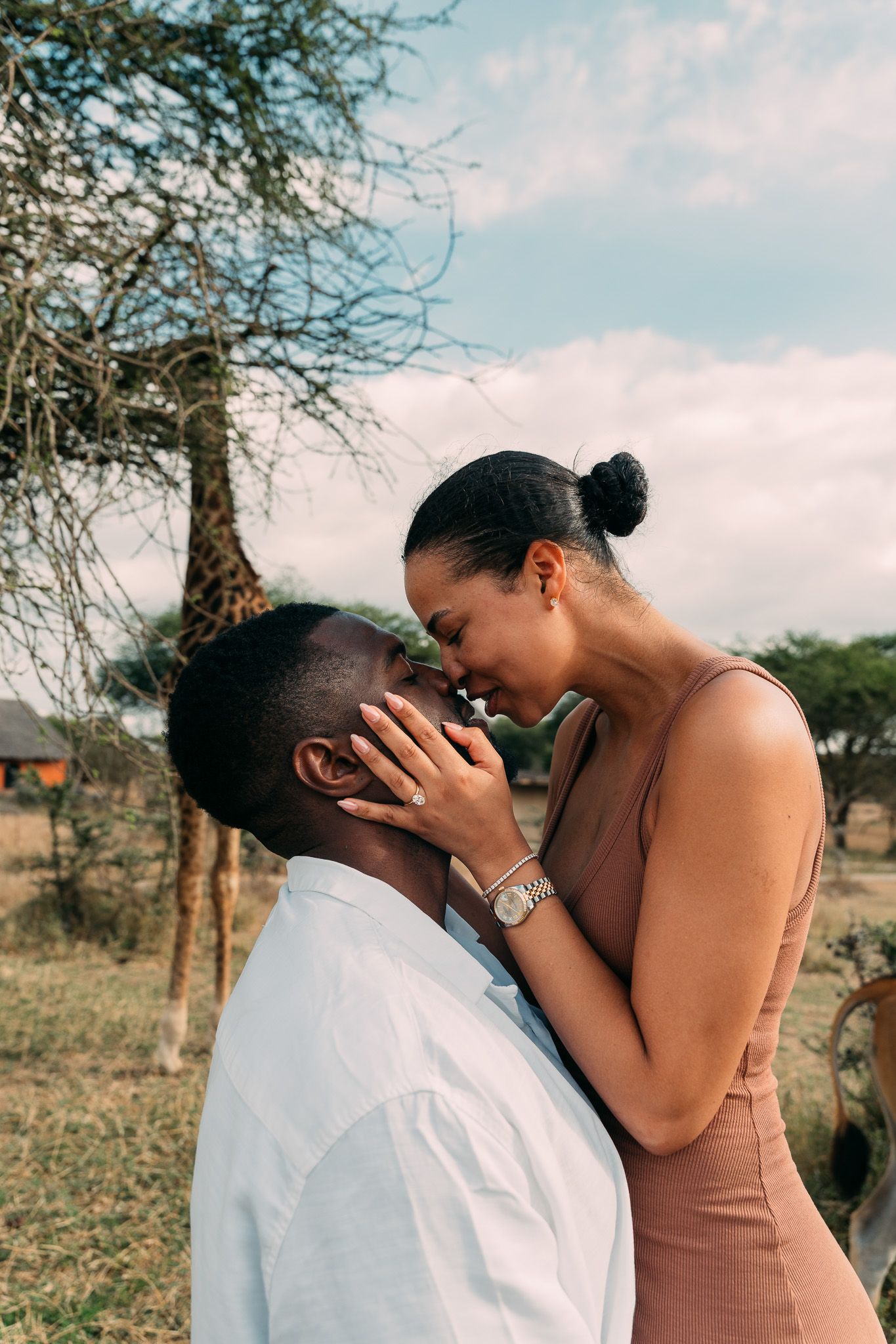 Couple nose-to-nose with engagement ring visible — giraffe standing near tree in background at Serval Wildlife Park