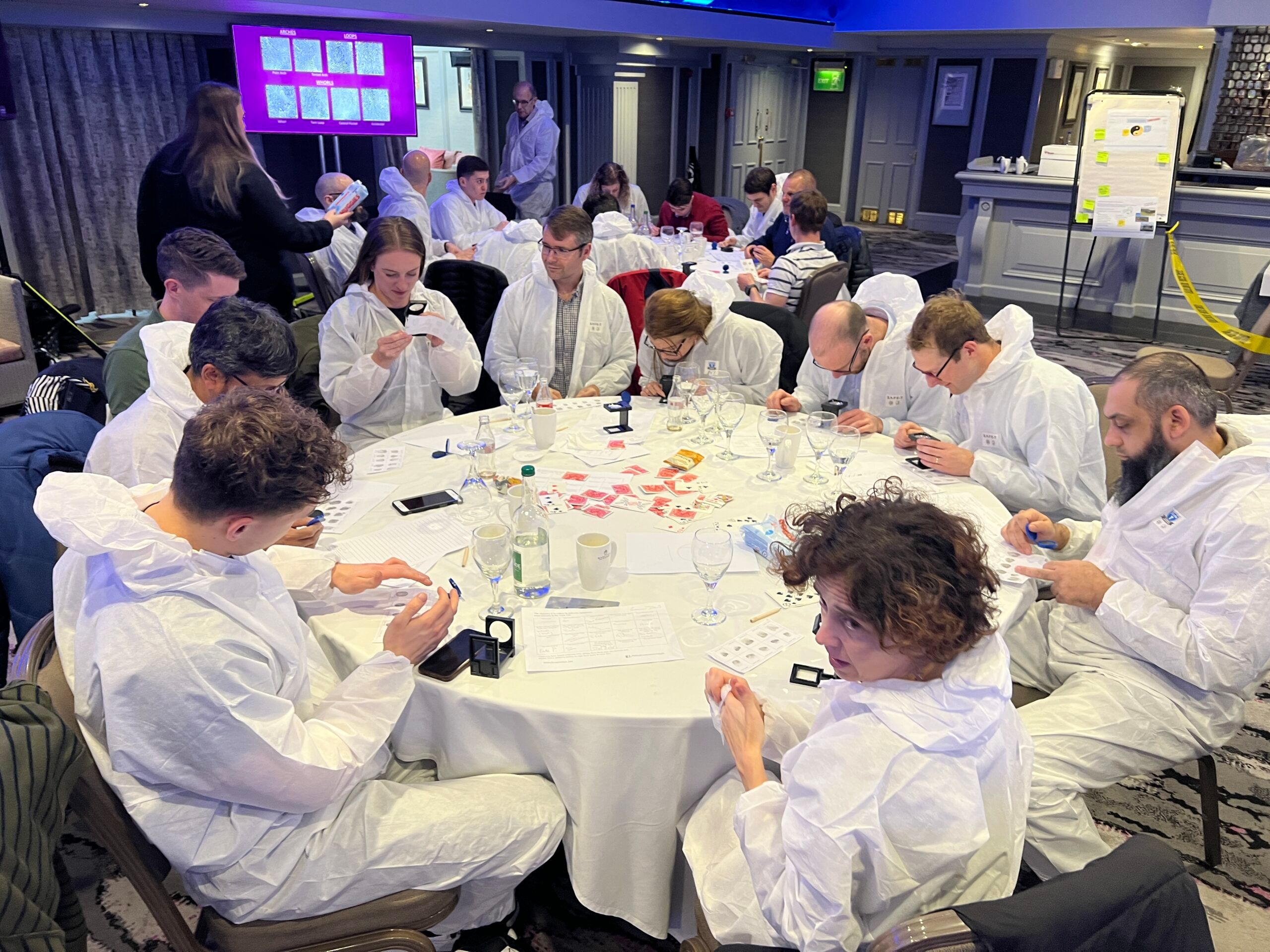Group of people in white coveralls examining small objects and cards around a round table during a Crime Scene Investigation team building event by Event Collection