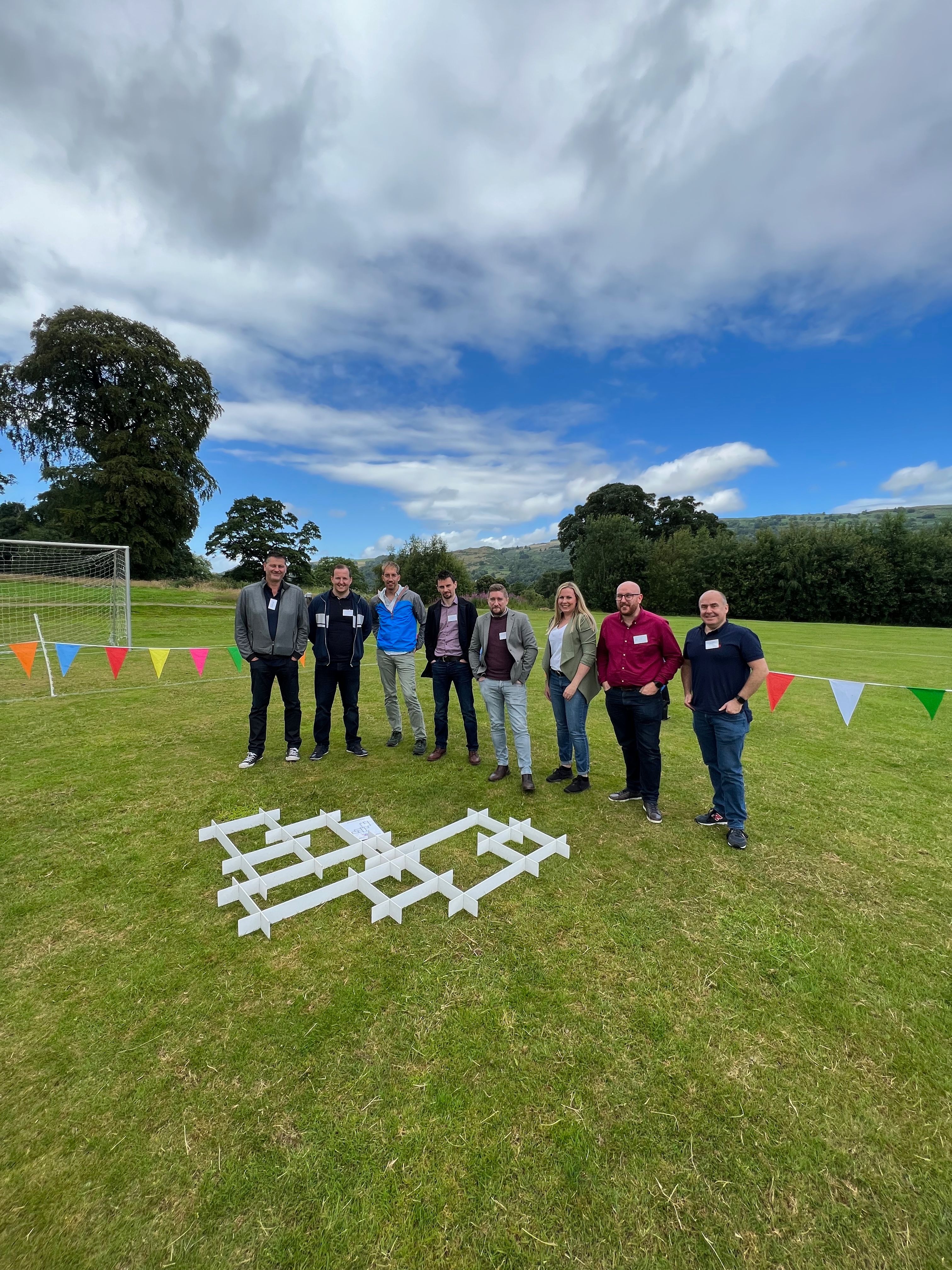 Take Me Back team building event — eight people standing behind a white grid structure outdoors with colourful bunting and blue sky