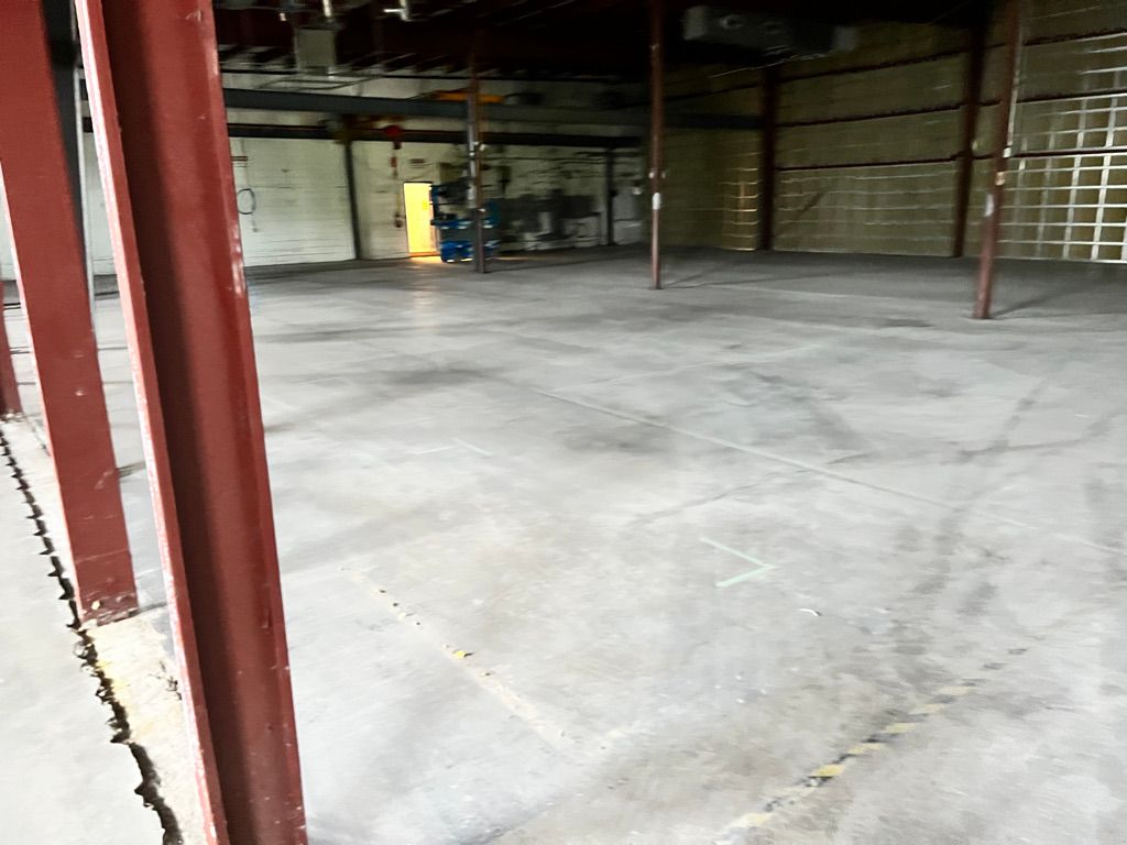 Large empty industrial space with concrete floor, reddish-brown metal support beams and brightly lit doorway in background