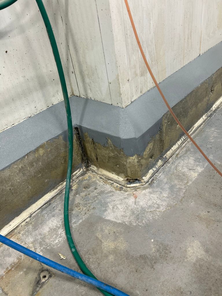 Wall-to-floor corner with polymer cove base in progress and colored utility hoses in commercial facility