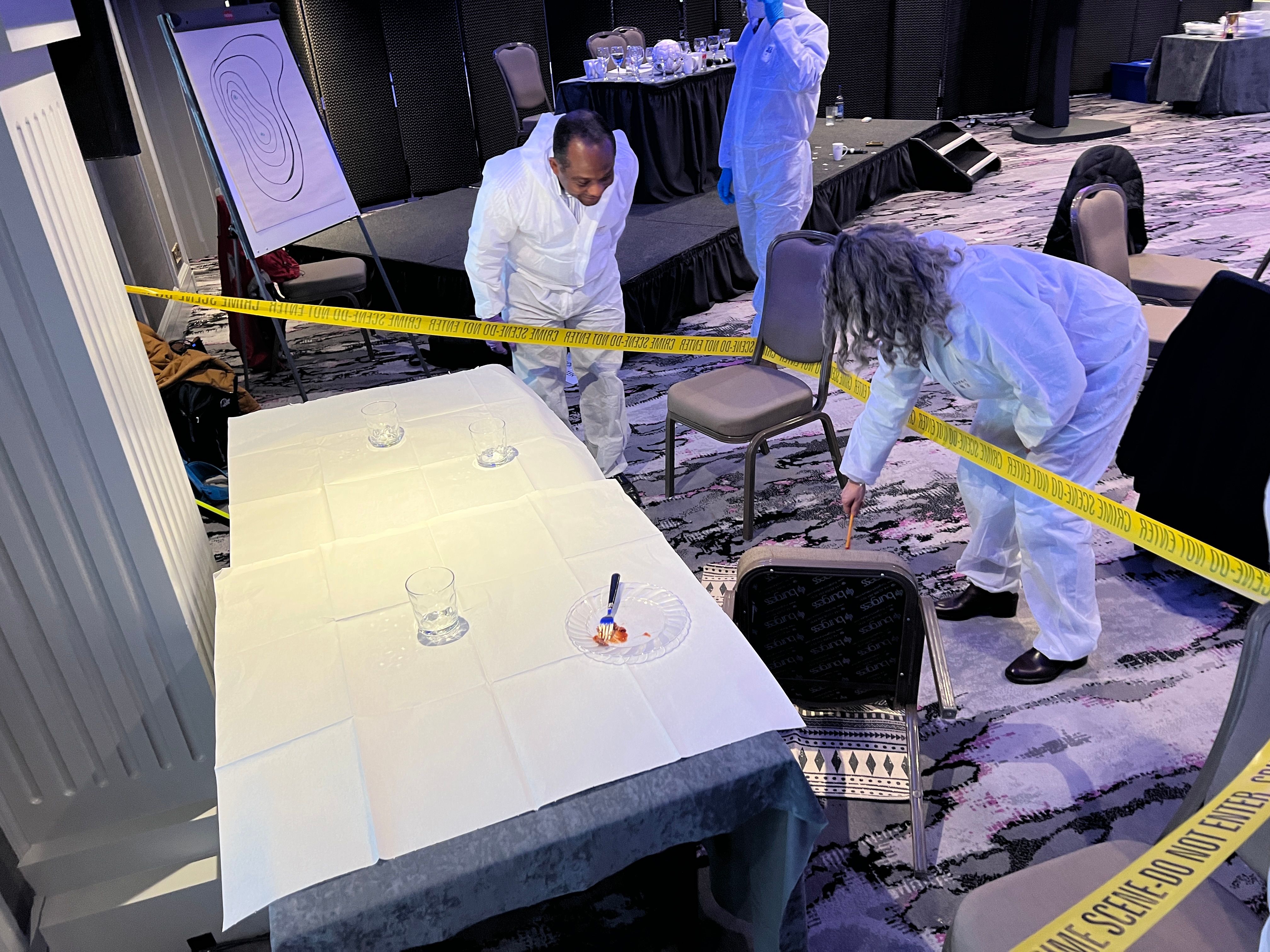 Three people in white forensic suits and blue gloves examining a crime scene with yellow caution tape and overturned chair