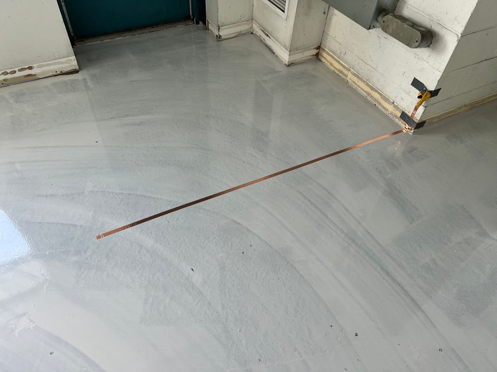 Room corner with smooth shiny gray ESD floor, copper grounding strip running diagonally to wall baseboard — teal door in background