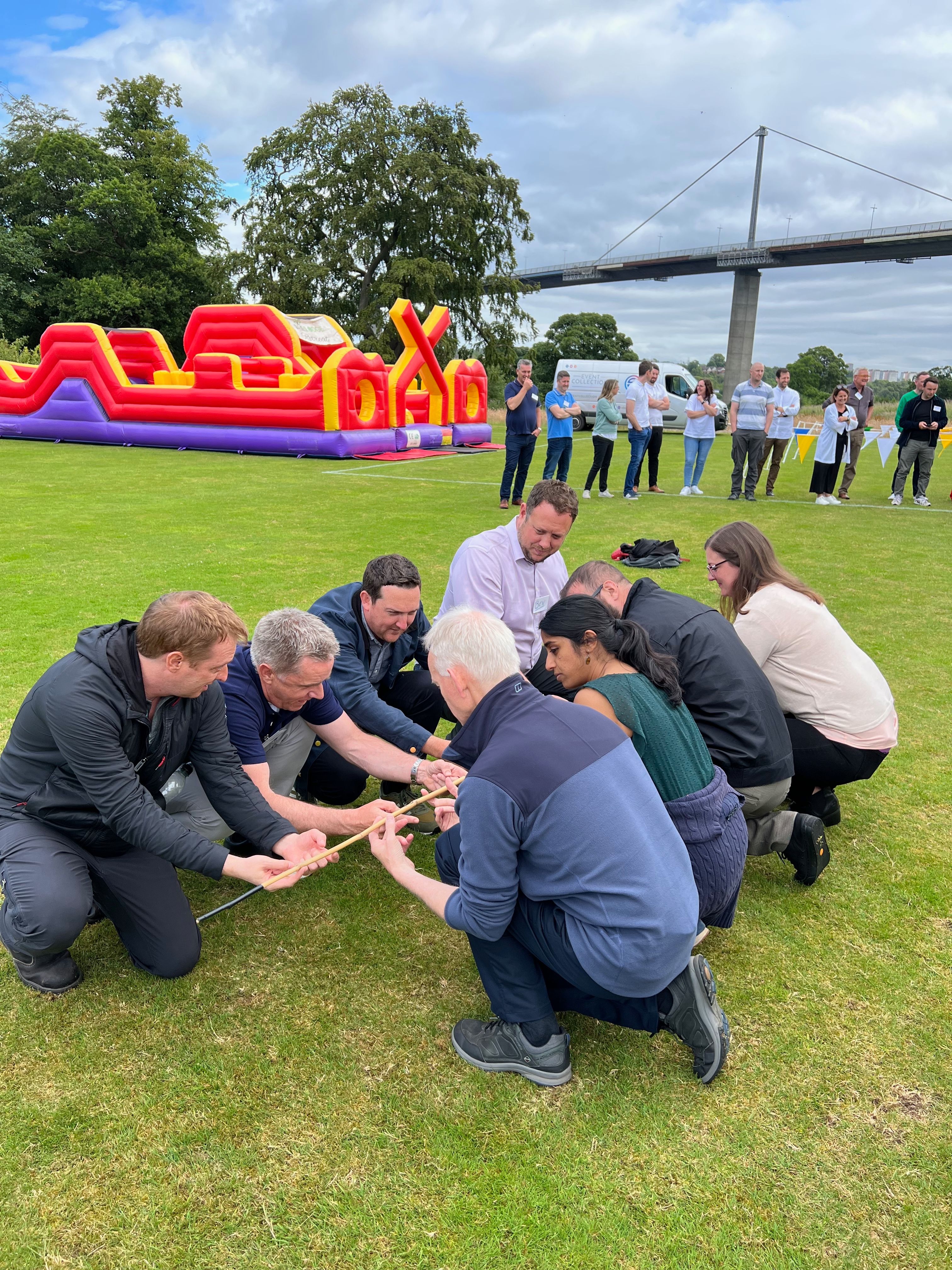 Take Me Back corporate team building — eight colleagues kneeling on grass holding a long stick together with an inflatable obstacle course in the background