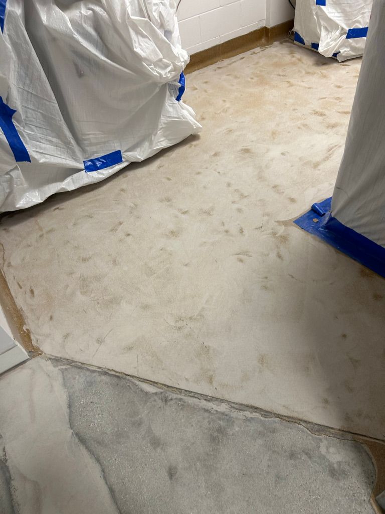 Pharmaceutical room during floor preparation — old material partially removed, exposed concrete sub-floor visible with protected furniture