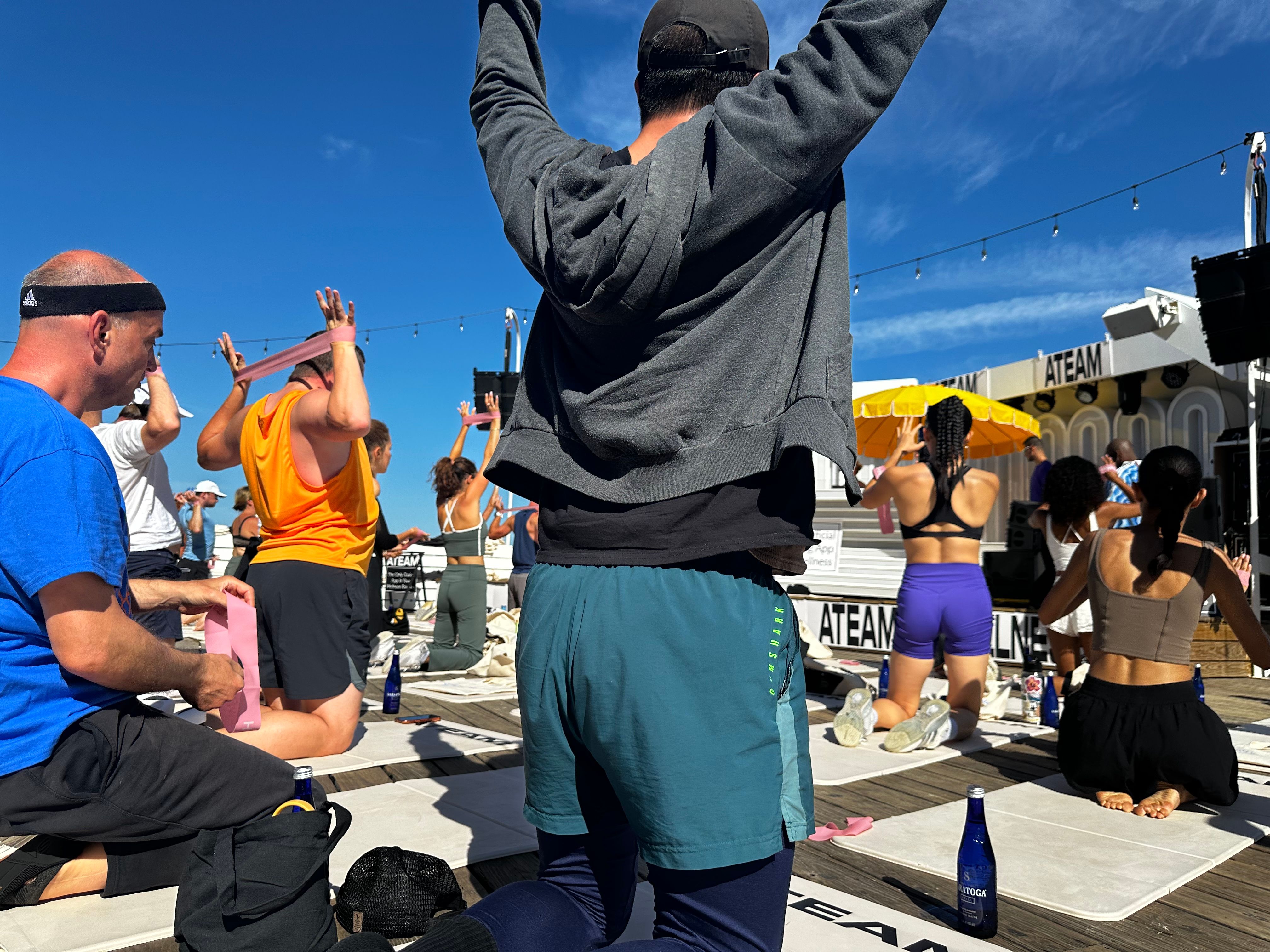 A-Team outdoor workout session on deck — group kneeling on mats with pink resistance bands, ATEAM sign in background, clear blue sky