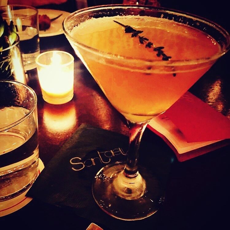 Santaella cocktail — golden martini with herb garnish on a Santaella-branded napkin, candlelight, warm speakeasy atmosphere