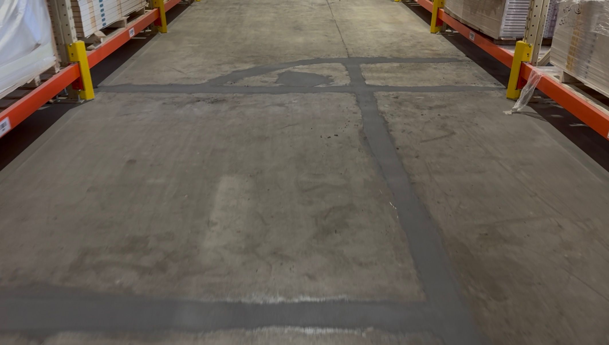 Warehouse aisle before-shot showing patchwork concrete floor with orange and yellow shelves and boxed goods