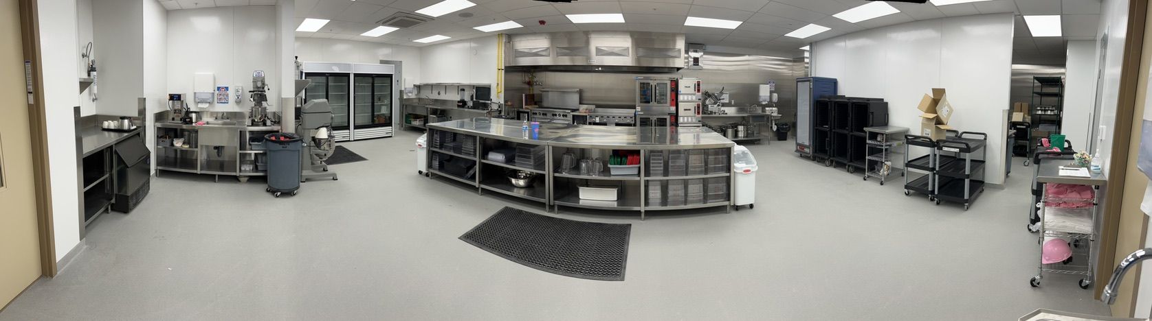 Large organized healthcare kitchen with stainless steel counters and shelves, refrigerators, black floor mats and supply carts — seamless light gray floor