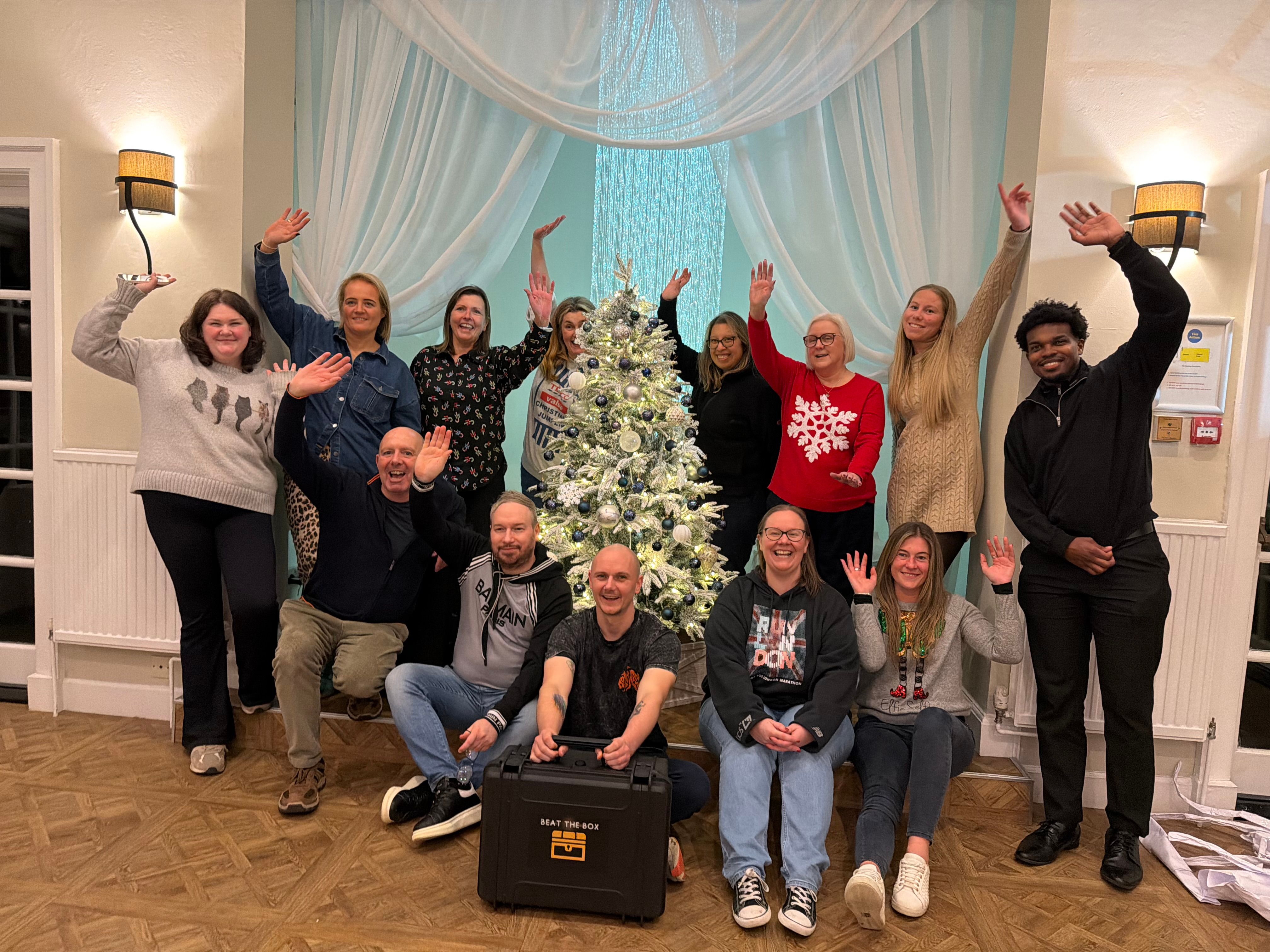 Beat the Box team building event — group of 13 celebrating with hands raised in front of a Christmas tree, holding the Beat the Box case