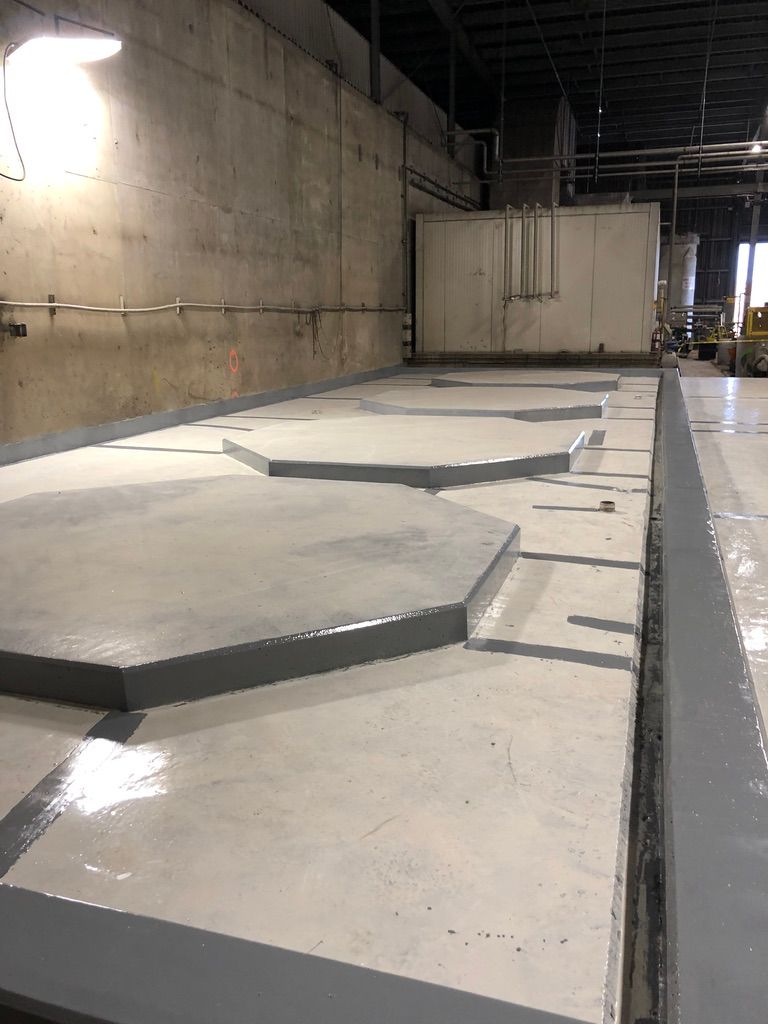 Industrial water or wastewater facility interior with seamless grey floor coating and raised hexagonal containment platforms, exposed conduit and pipes along the walls