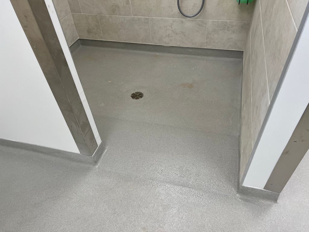 Floor drain detail in a pharmaceutical facility — textured seamless gray floor with circular stainless steel drain cover and coved tile wall base
