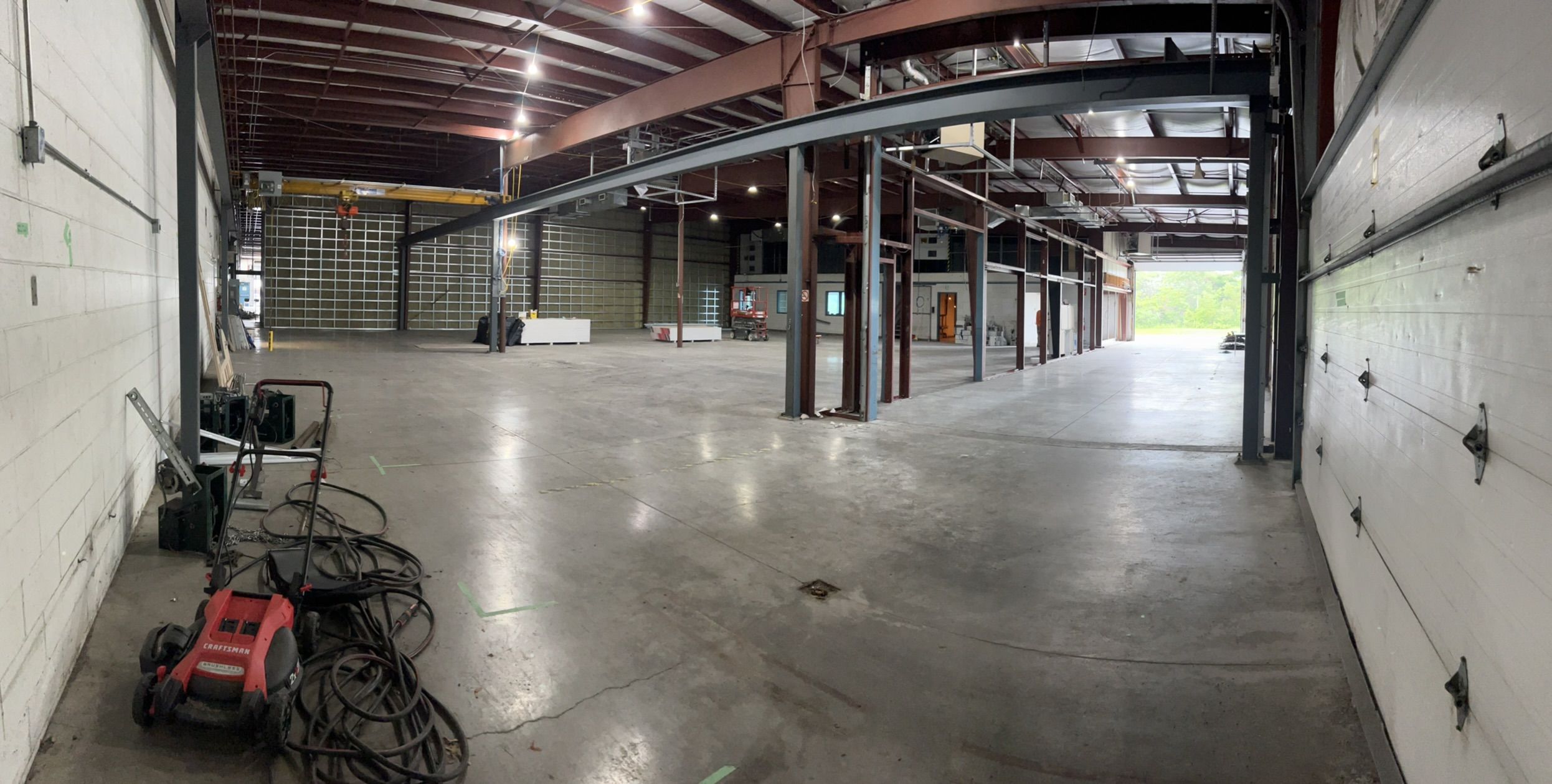 Large mostly empty industrial warehouse with concrete floor, red lawn mower and white garage door — open door revealing exterior greenery