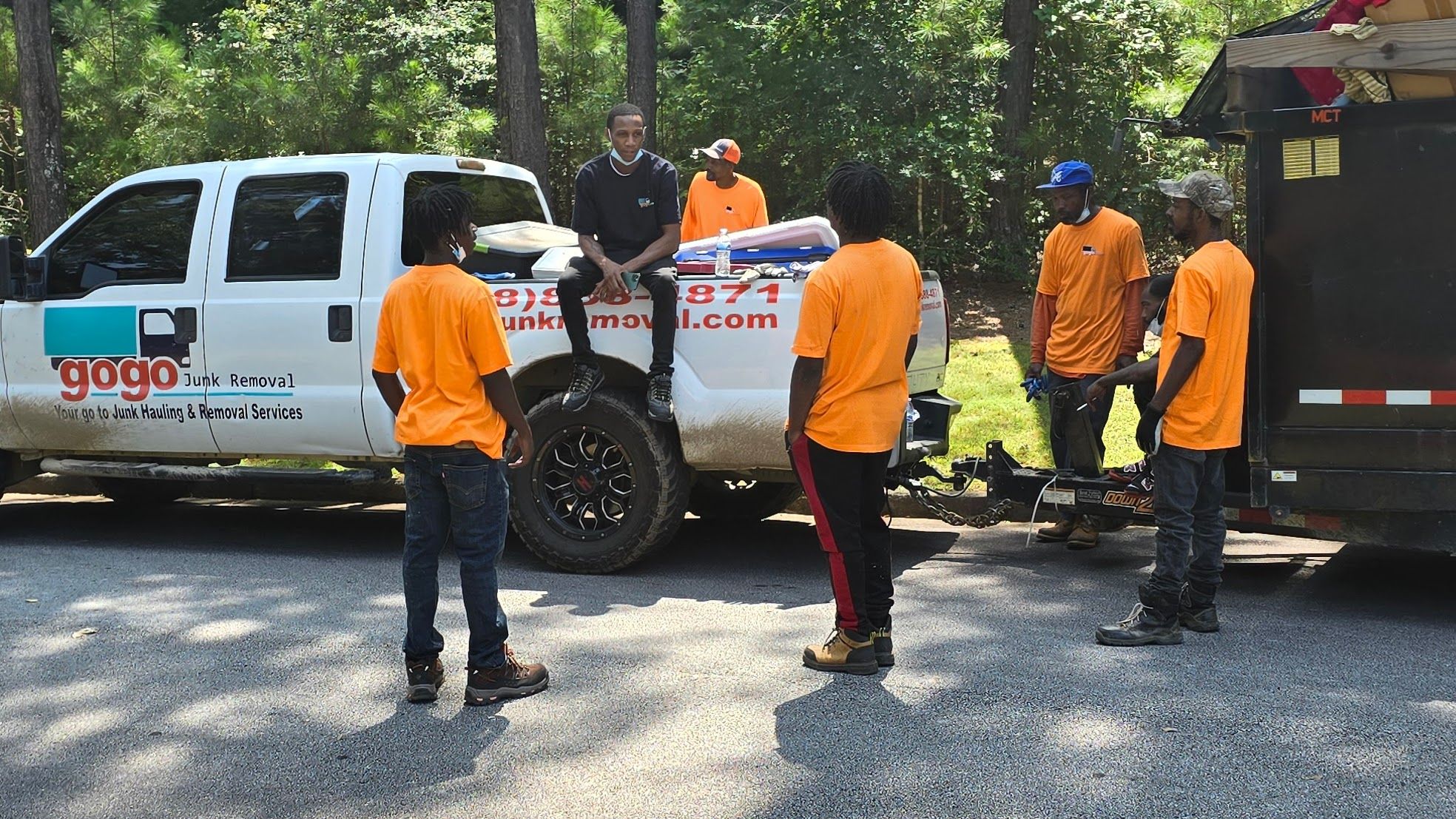 GoGo Junk Removal full team of six people in branded orange and navy shirts posing near white truck and black trailer