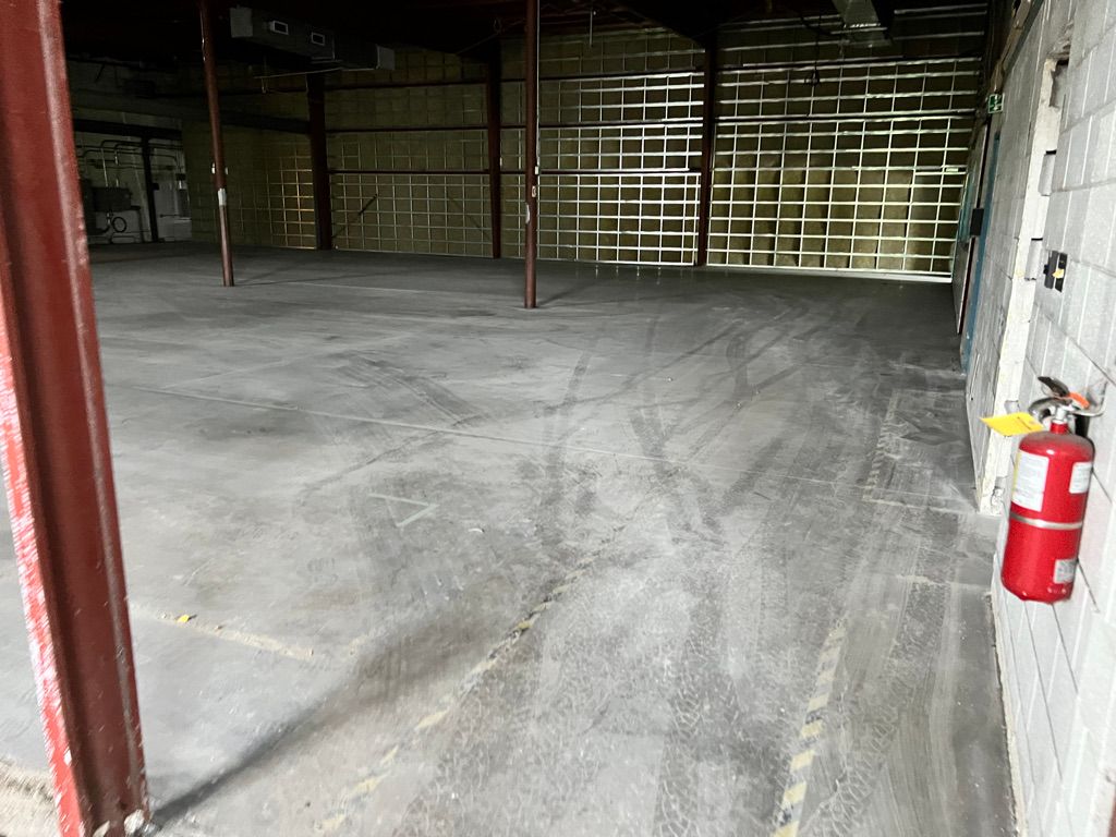 Empty industrial warehouse with concrete floor showing tire marks, red metal beams and mounted fire extinguisher on block wall