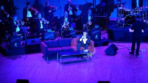 Drew Anthony as Dean Martin seated on red couch with live orchestra
