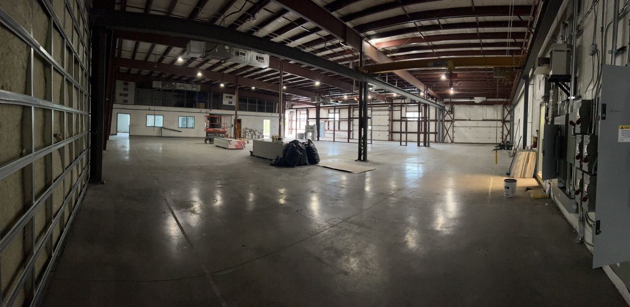 Large empty industrial warehouse with concrete floor, exposed steel beams, multiple garage doors and construction materials near center