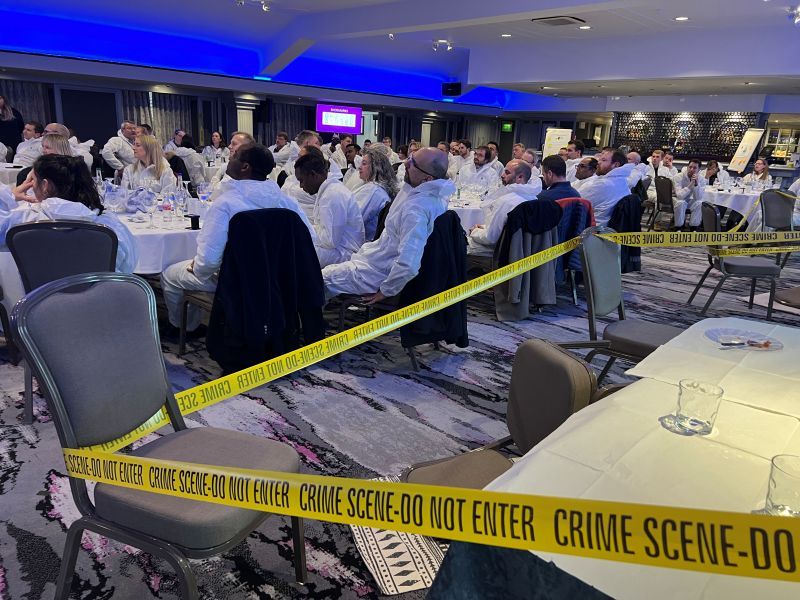 Large group of people in white protective suits seated at round tables with yellow crime scene tape across the room