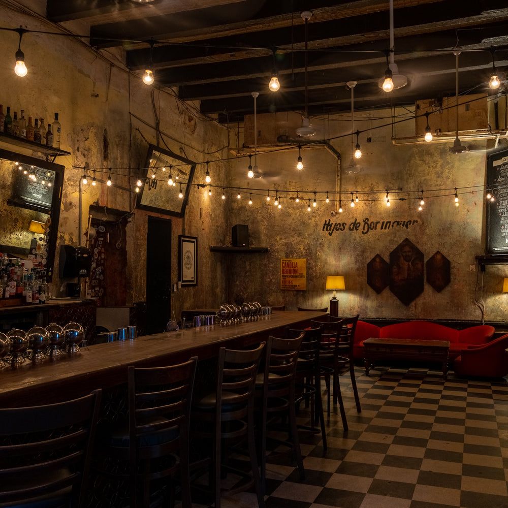 La Factoría Front Bar — rustic run-down chic interior with long wooden bar counter, string lights, checkered floor, red couch, Hijos de Borinquen wall art, and warm ambient lighting in Old San Juan