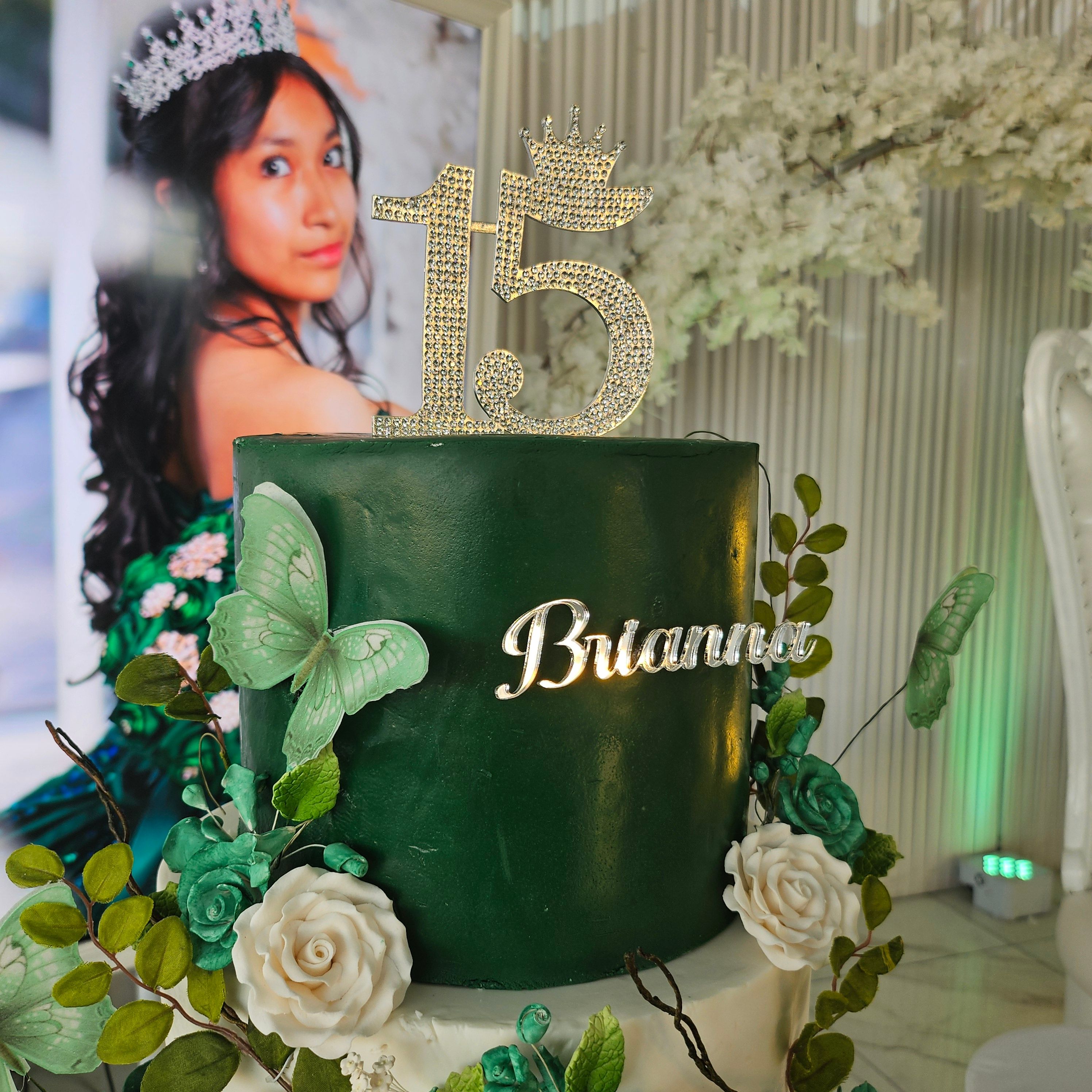 Grand tiered quinceañera cake with green and white floral design, crown topper, and personalized name plaque by NYCakes Boutique