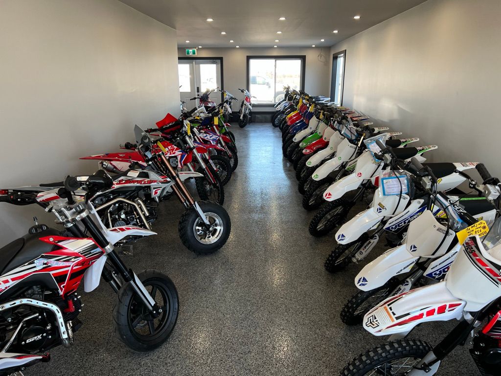 Narrow retail showroom with smooth gray seamless floor and two rows of red and white dirt bikes facing a glass door