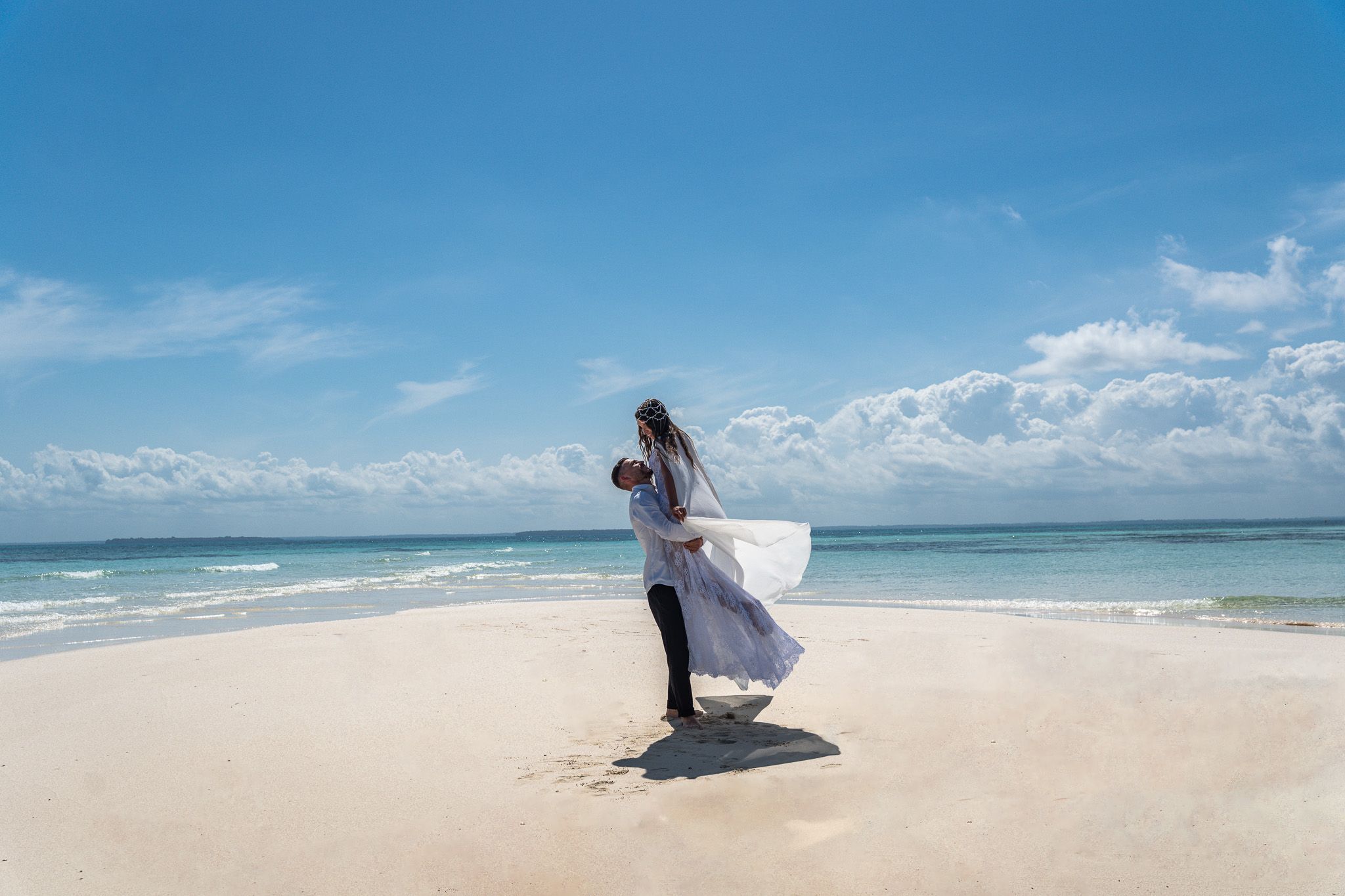 Man lifting woman on white sand beach with turquoise ocean backdrop