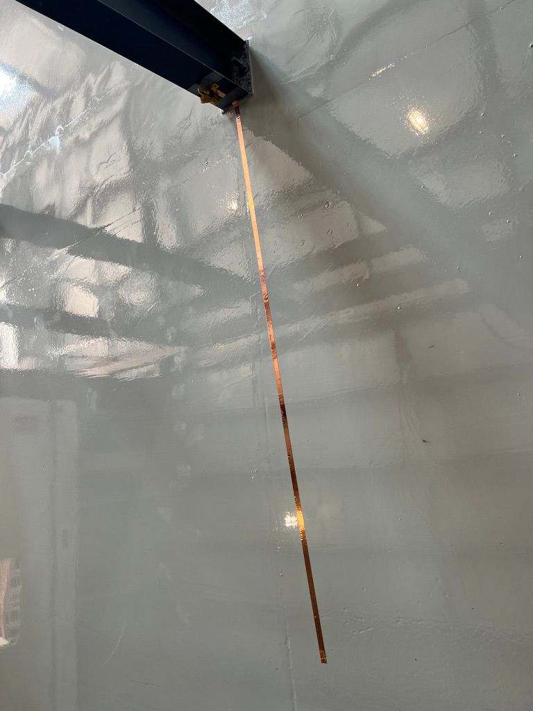 Shiny reflective copper ESD grounding strip hanging vertically from dark horizontal beam on smooth glossy gray wall