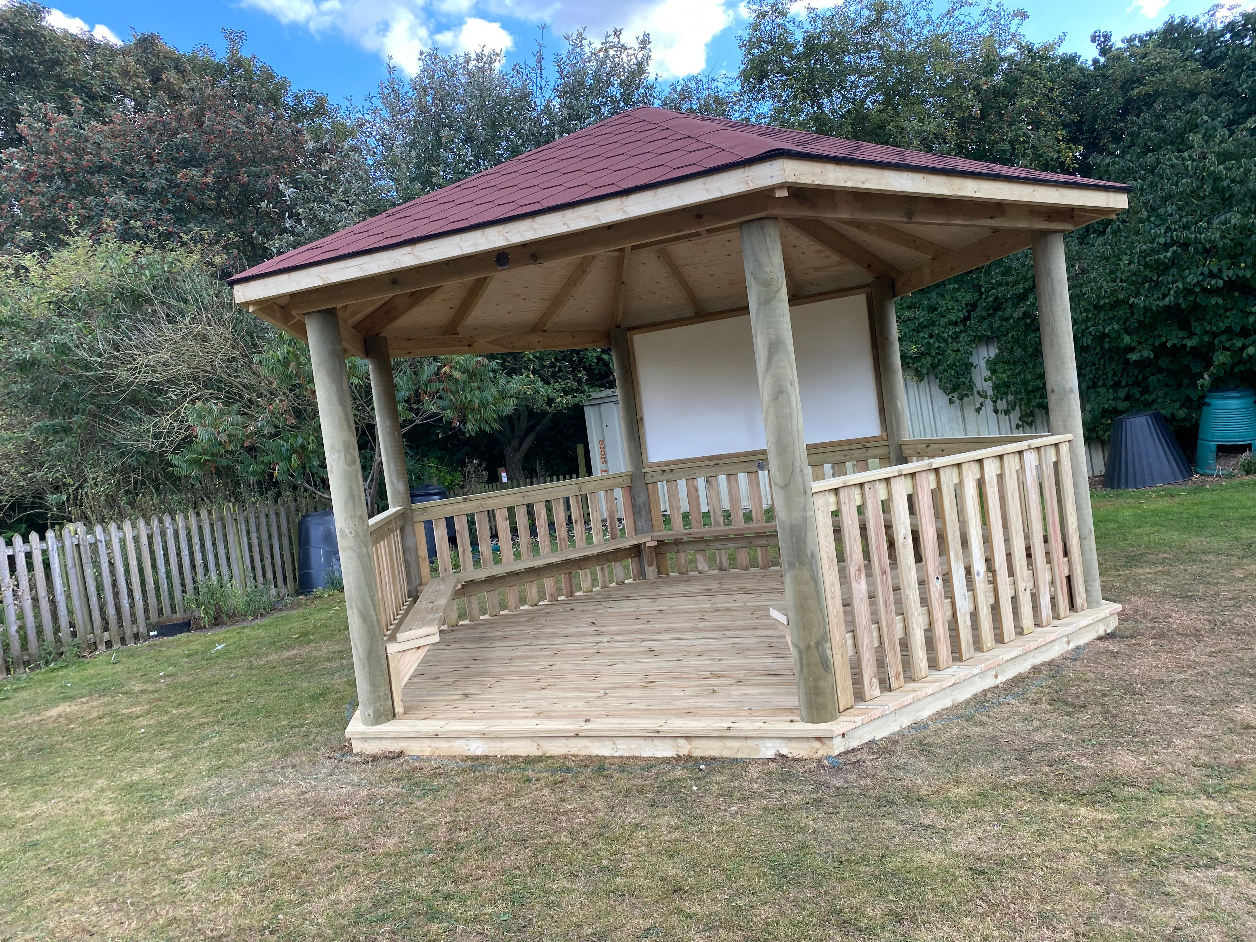 Timber hexagonal gazebo with felt shingle roof, open sides and bench seating on a grassy school site
