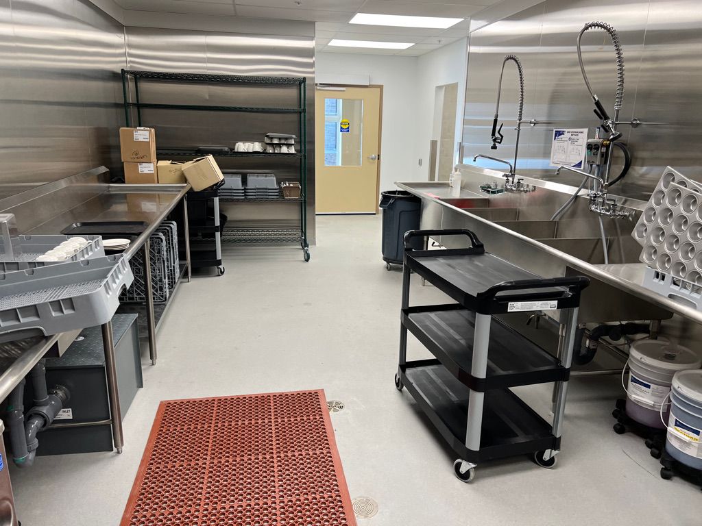 Clean healthcare kitchen with stainless steel countertops, triple sink with spray faucets and shelving — seamless light-colored floor with anti-slip mat