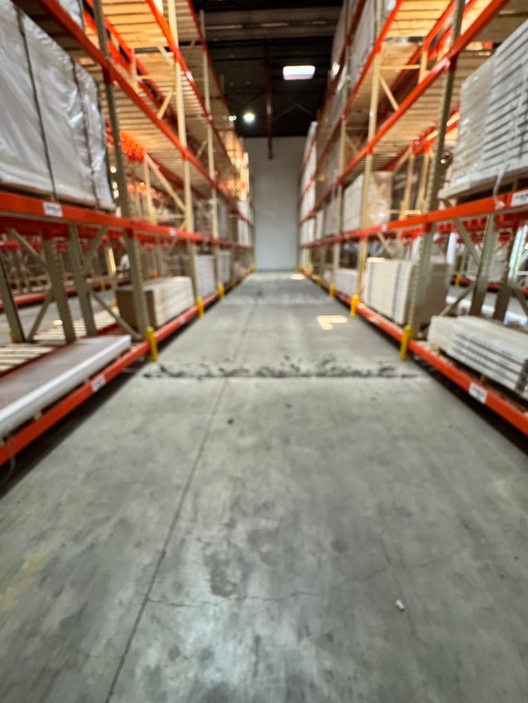Warehouse aisle before-shot showing rough cracked concrete floor between metal shelving with palletized product
