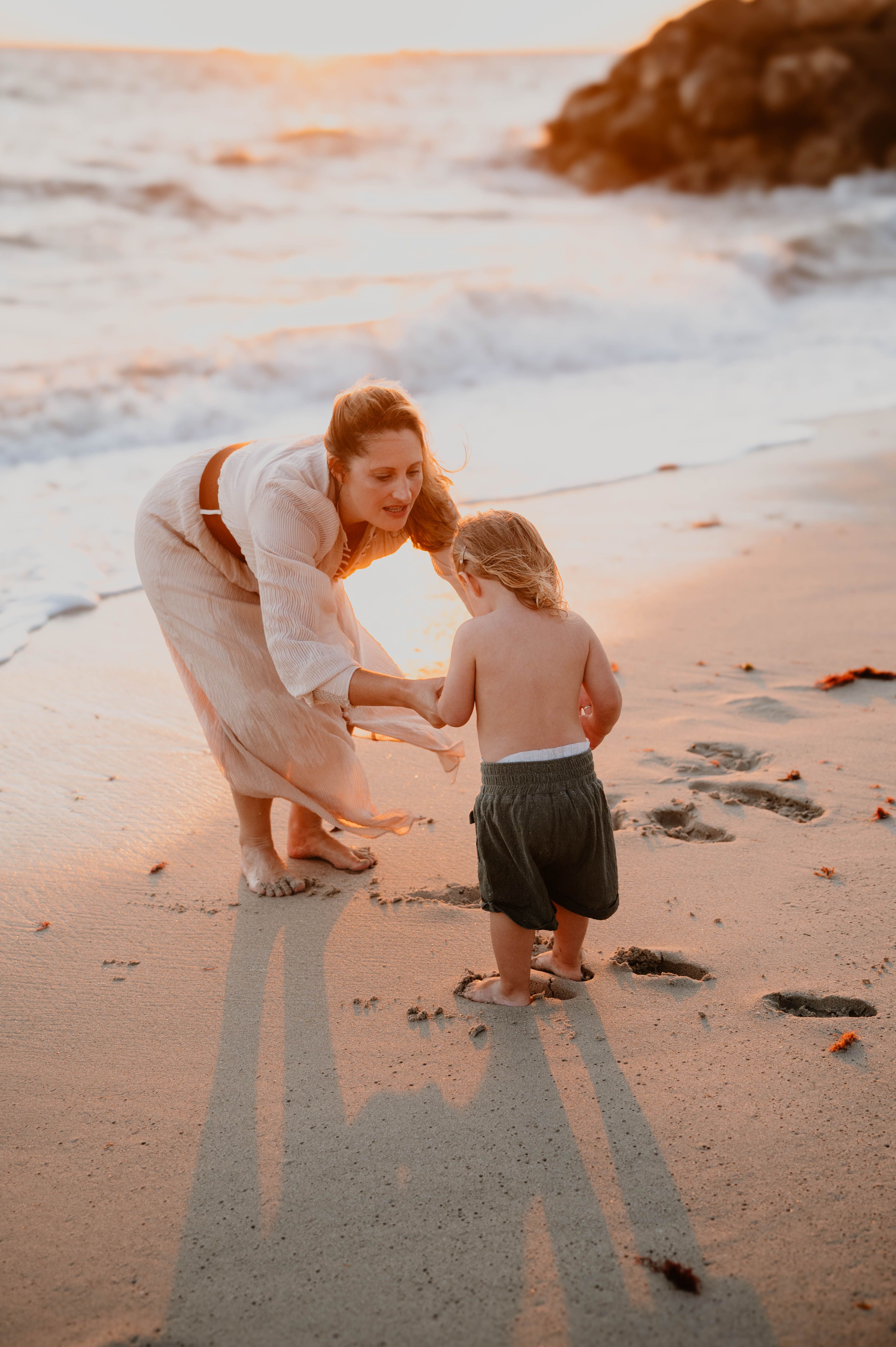 Parent holding a young child's hands on the beach at sunset