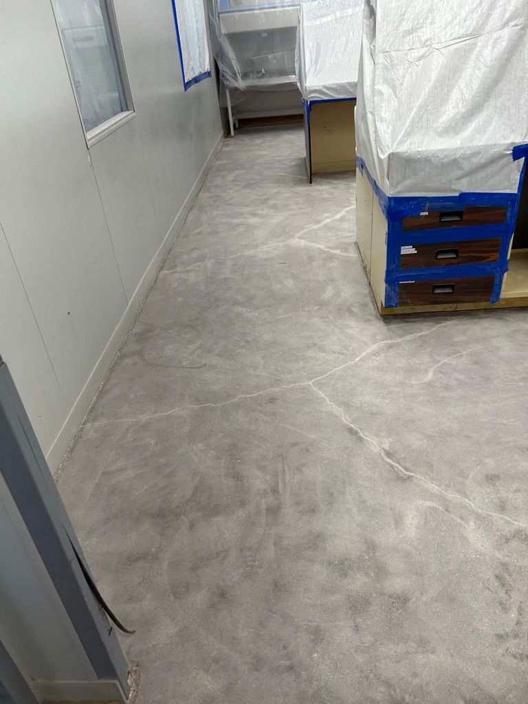 Narrow pharmaceutical facility room with cracked concrete floor, protected equipment and natural window light prior to epoxy resurfacing