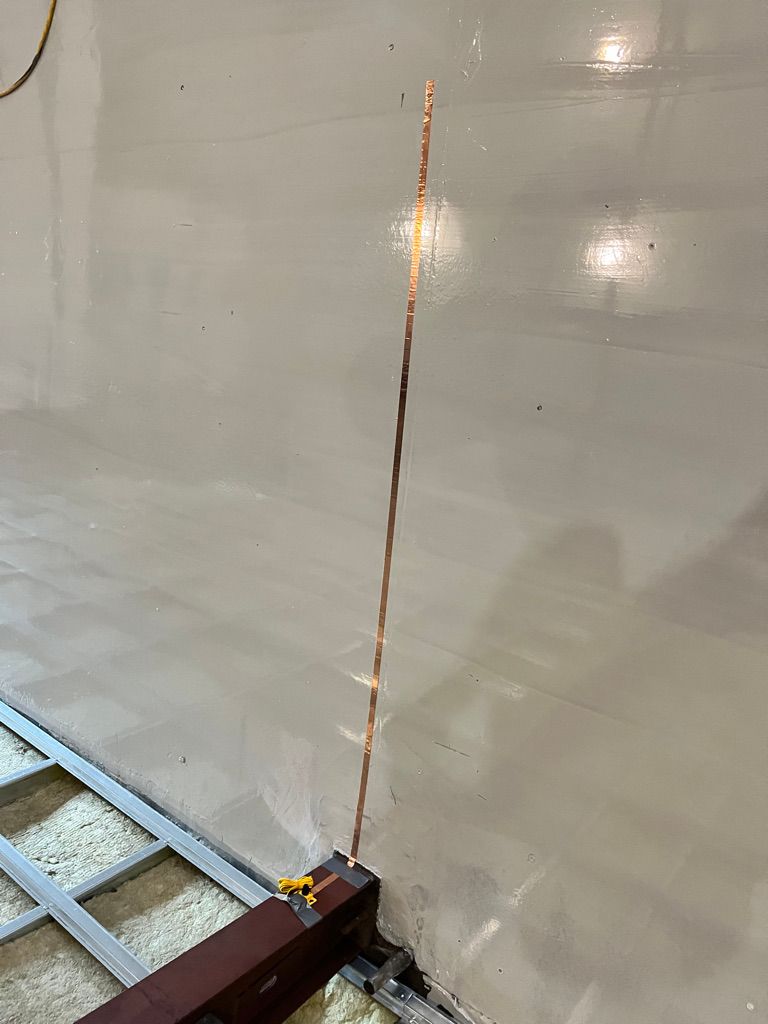 Close-up detail of wall-mounted vertical copper grounding strip with yellow wire on beam and metal floor framing with insulation
